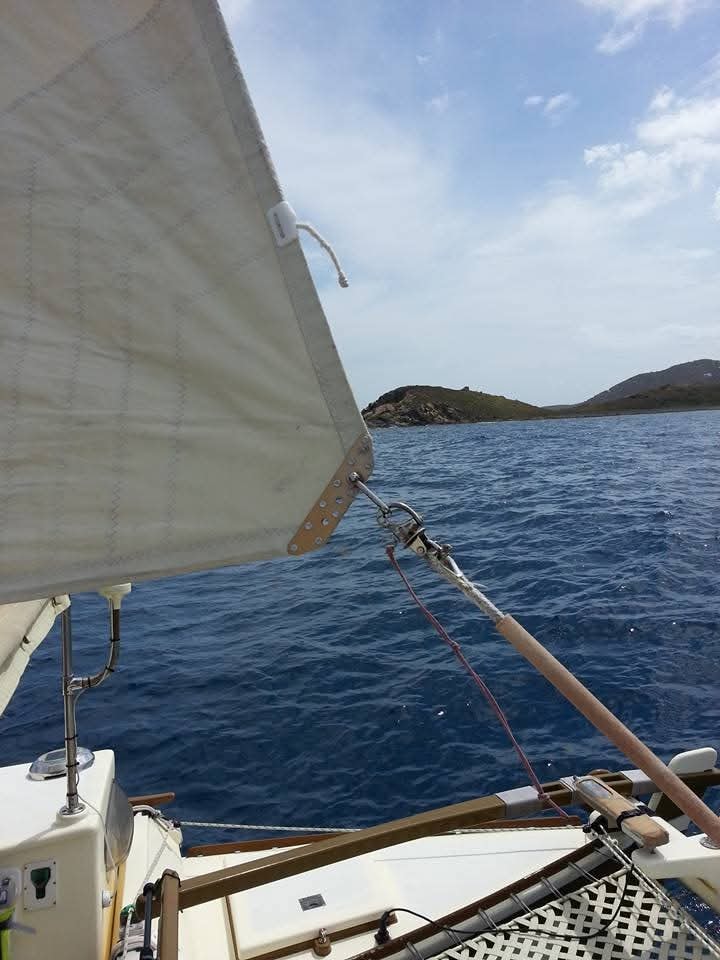 View from the Mahiya cockpit past the mainsail toward a lush green VI island on deep blue water under blue sky