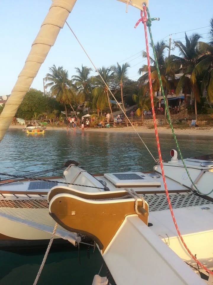 View from the Mahiya deck past the mast toward a tropical beach with palm trees and colorful waterfront buildings