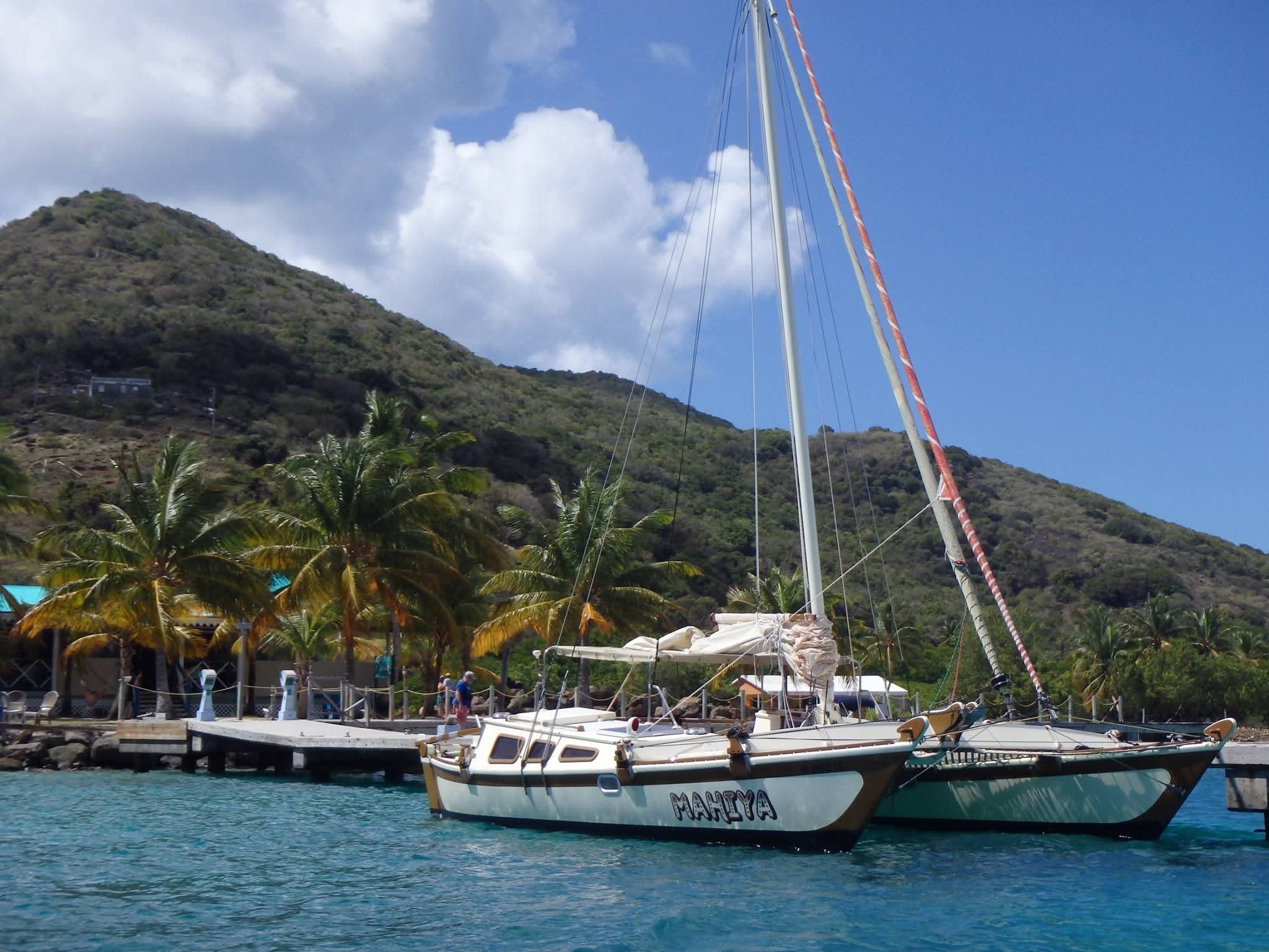 The Mahiya catamaran docked at a tropical pier with palm trees and a lush VI mountain behind, MAHIYA name on hull