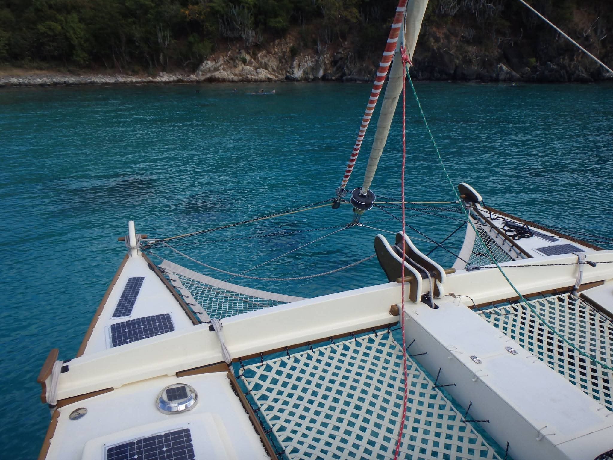View from the Mahiya bow over the lattice trampoline net toward turquoise water and a rocky VI coastline, solar panels visible