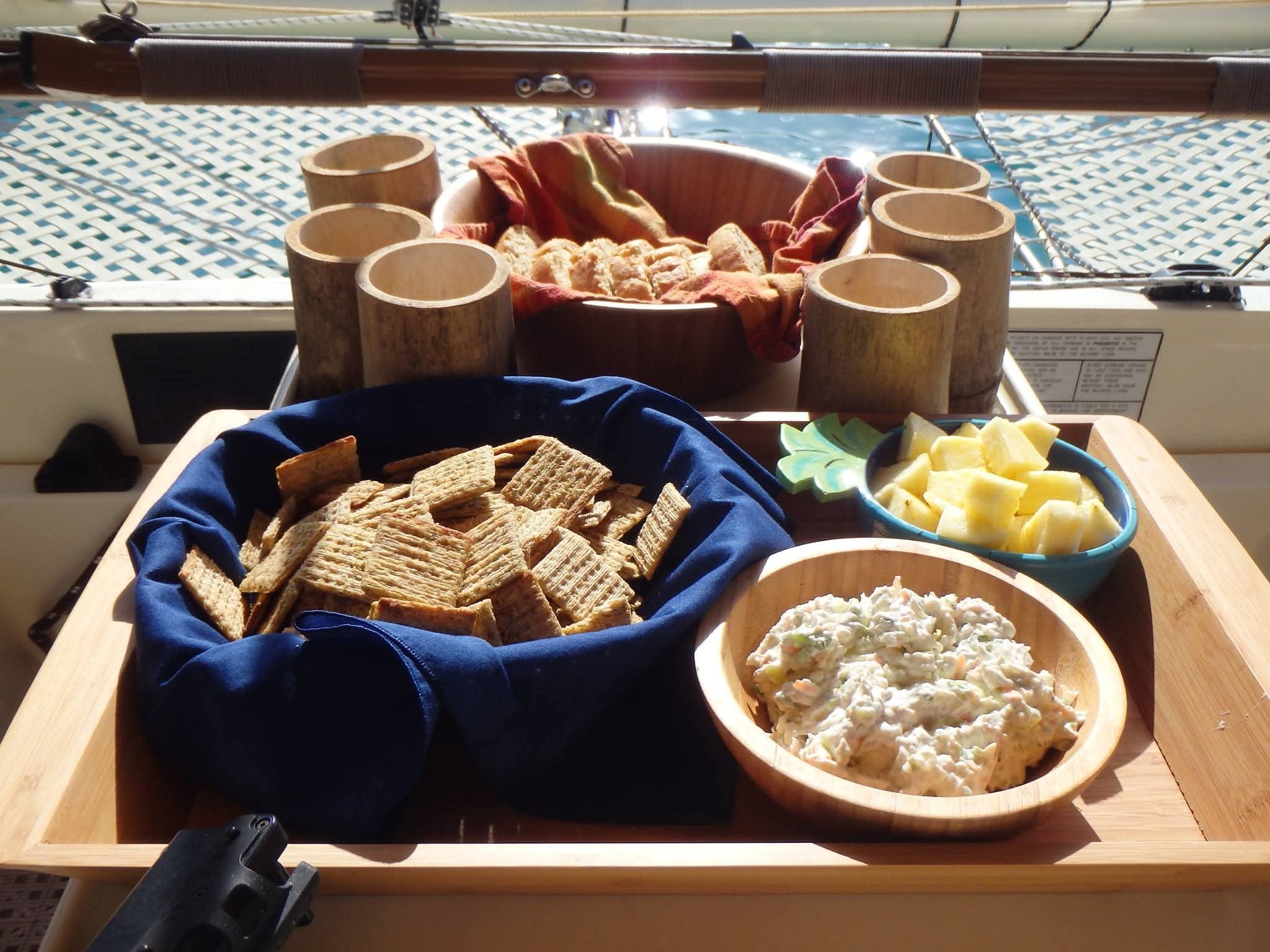 Crackers in a wooden bowl with chicken salad dip and fresh pineapple chunks served on the Mahiya deck, bamboo cups behind