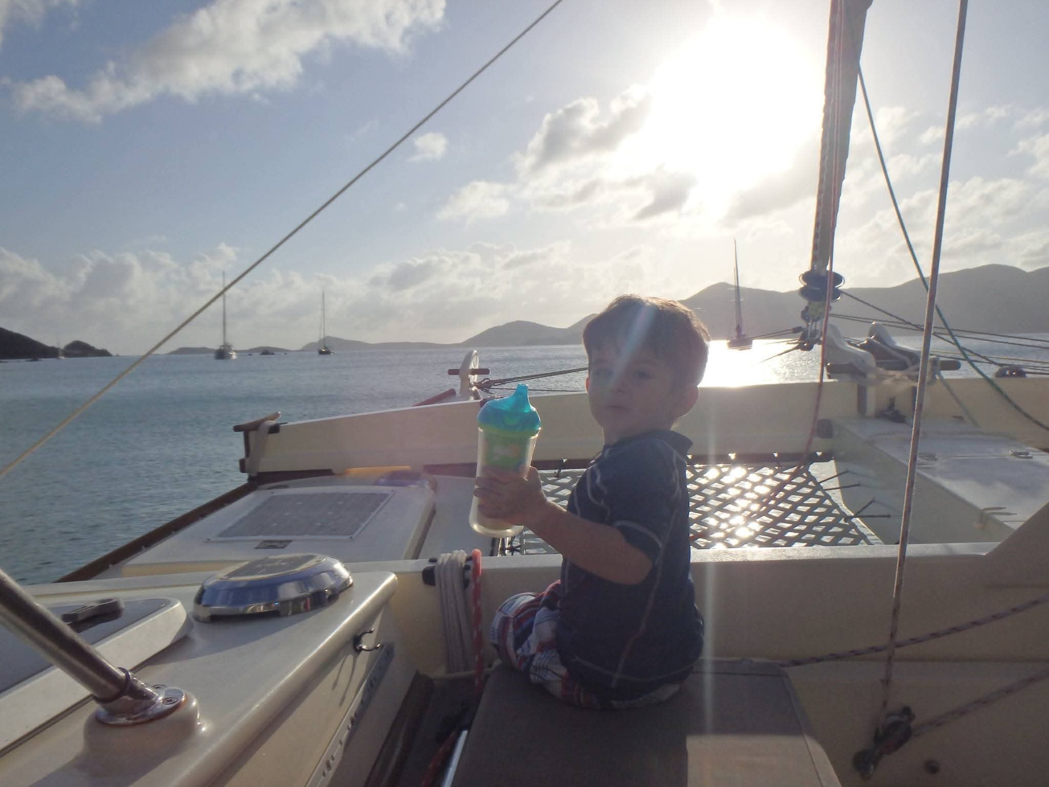 Young boy sitting on the Mahiya cockpit at dusk holding a sippy cup, VI mountain islands silhouetted behind