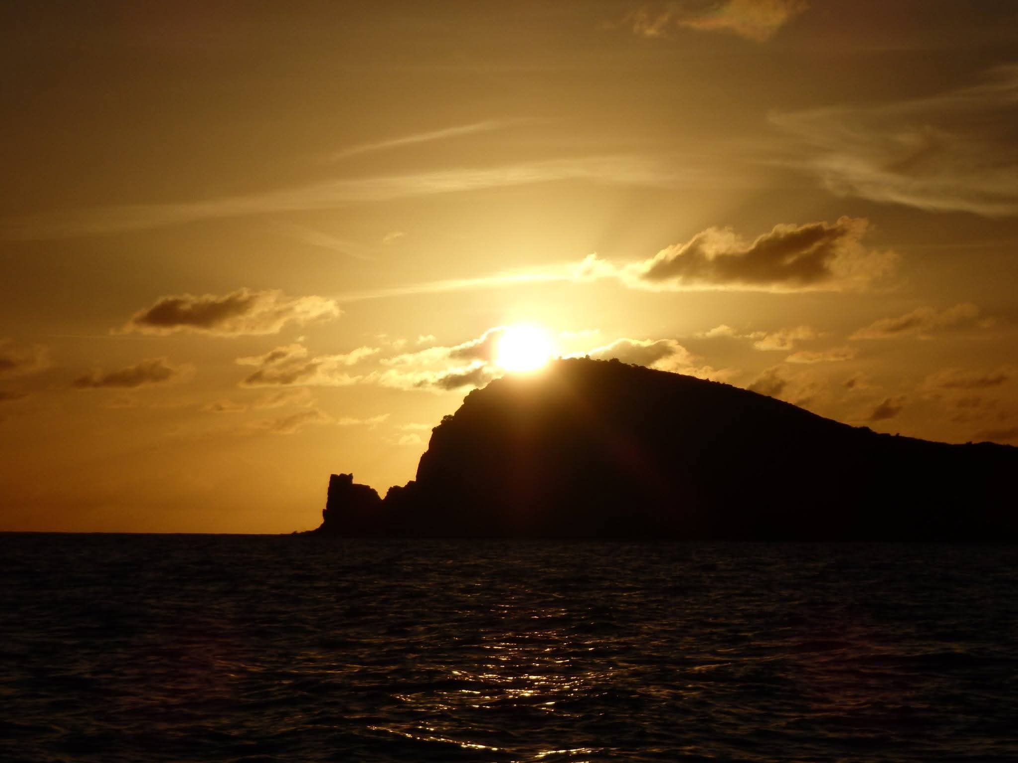 Dramatic golden sunset with the sun setting behind a dark rocky VI island silhouette over calm dark water