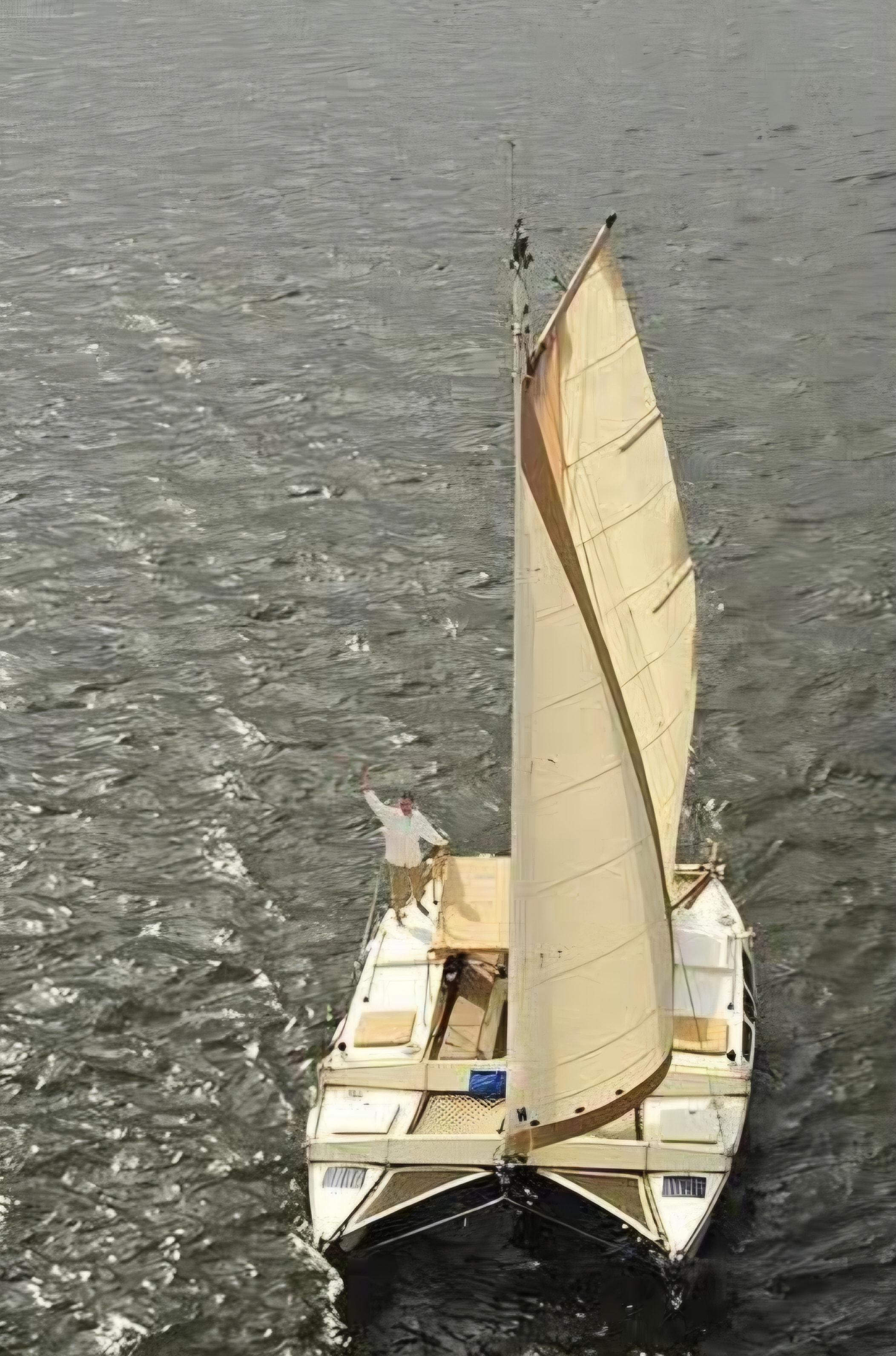 Aerial drone shot of the Mahiya catamaran under full sail on choppy grey water, person waving from the bow deck