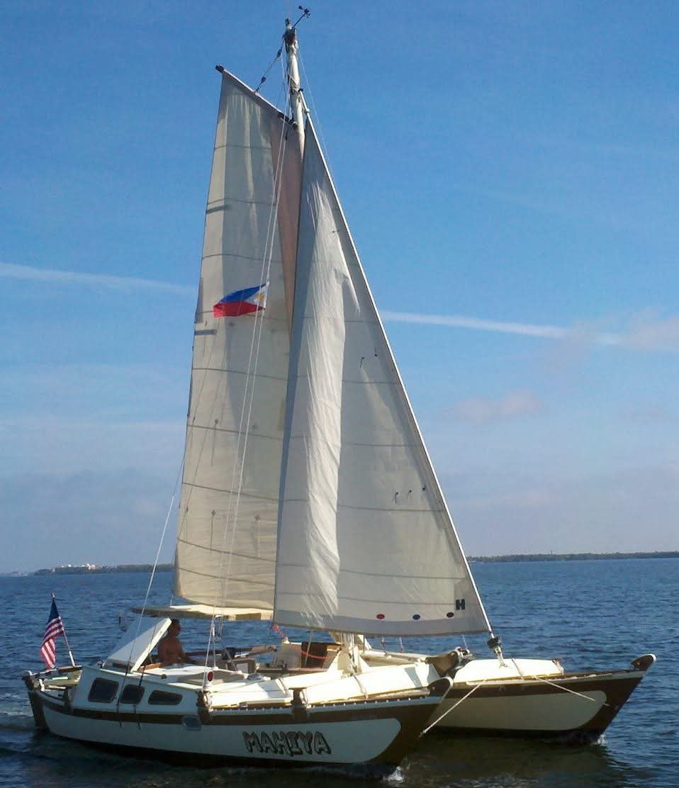 The Mahiya catamaran under full white sail with the Philippine flag flying and American flag at the stern, blue sky behind