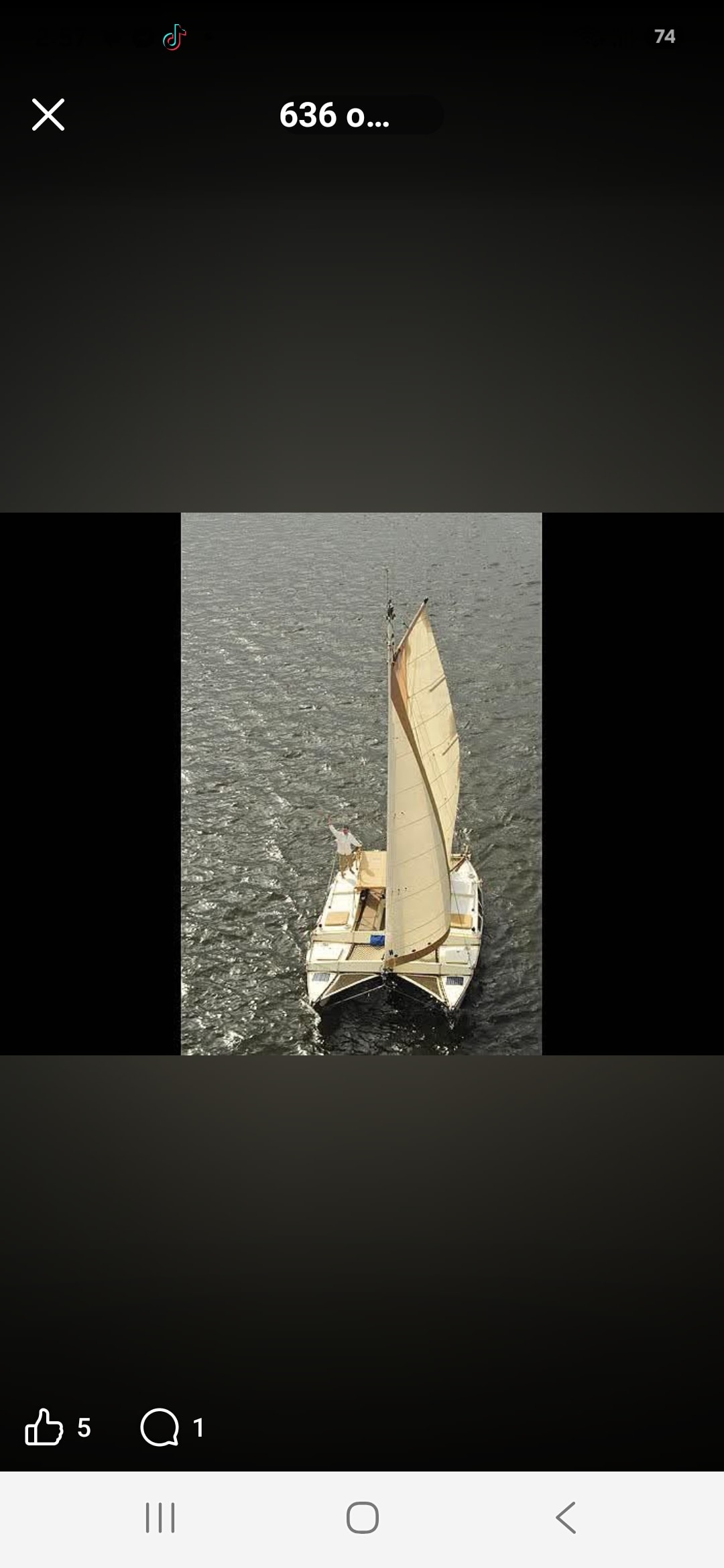 Aerial drone screenshot of the Mahiya under cream sail on choppy grey water, Captain Karl waving from the bow