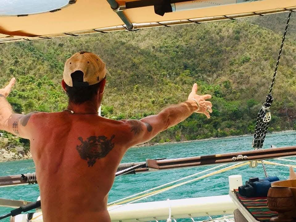 Captain Karl with arms outstretched on deck, lush green Virgin Islands hills behind