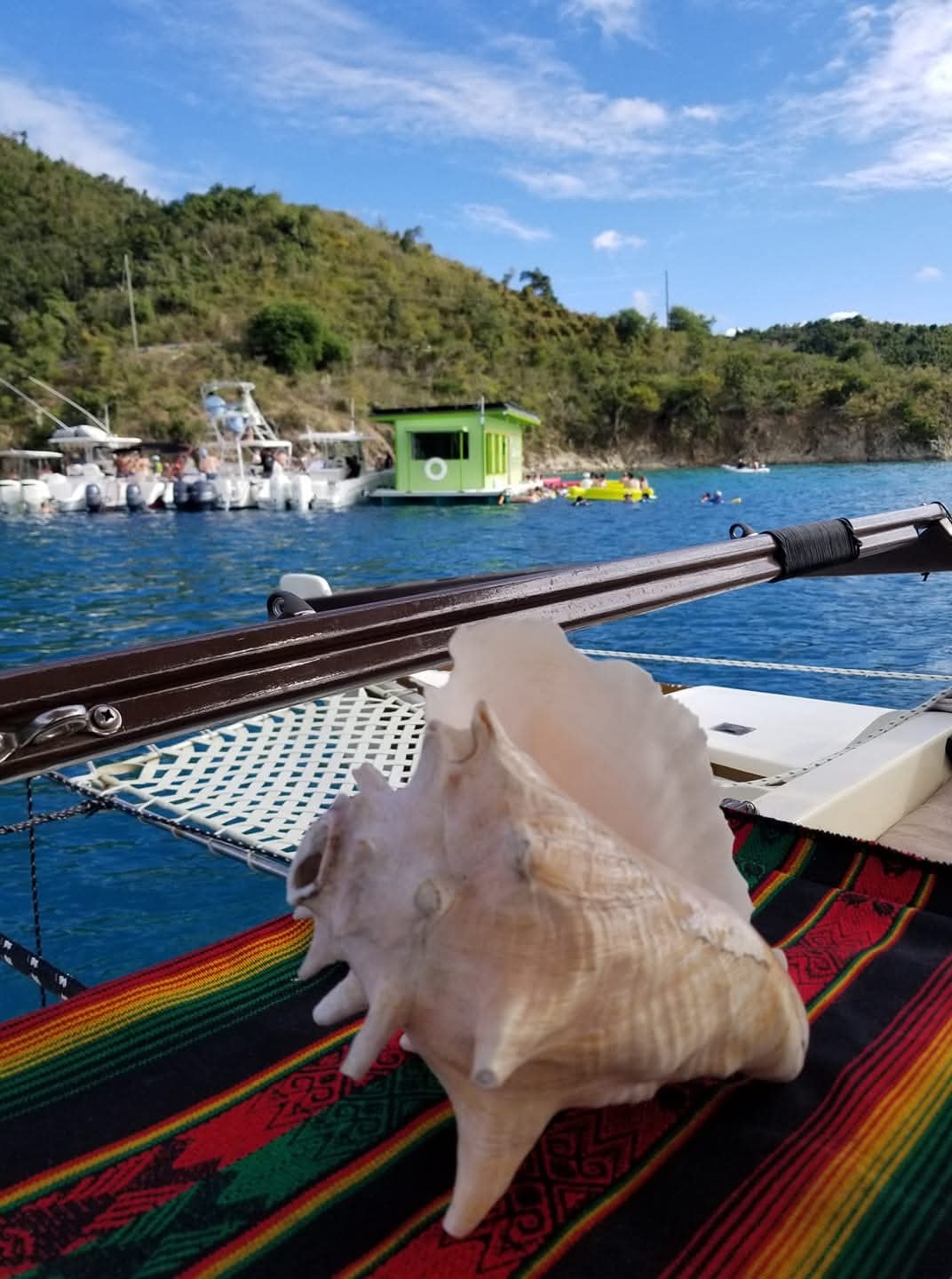 Conch shell on a colorful Rasta tablecloth on deck with a green boathouse cove behind