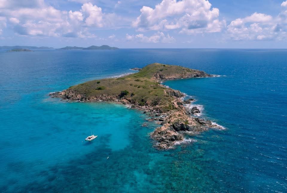 Aerial drone shot of a tiny rocky VI island surrounded by brilliant turquoise water, Mahiya anchored nearby
