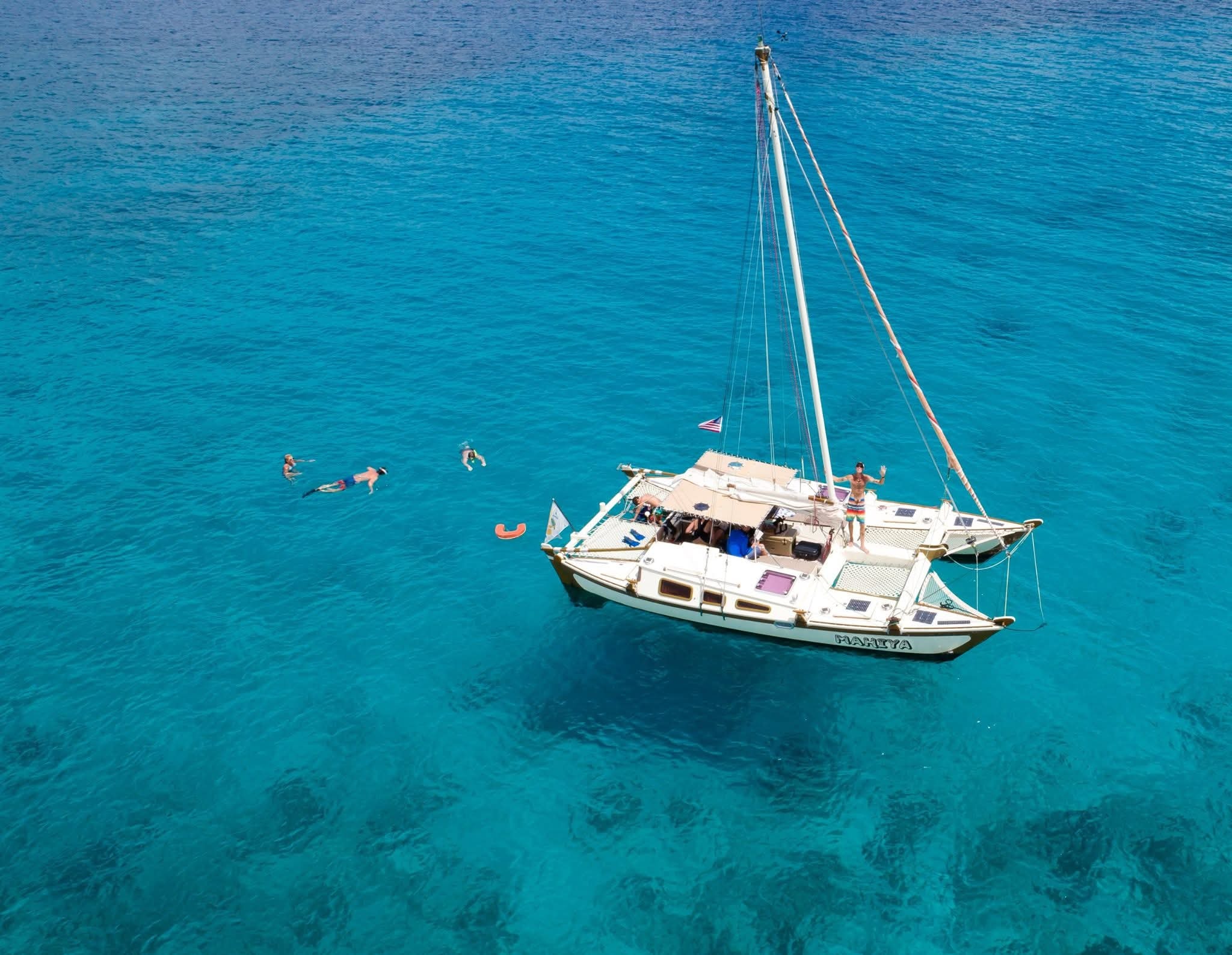 Aerial drone shot of the Mahiya anchored in crystal turquoise water with snorkelers swimming nearby, Captain Karl waving