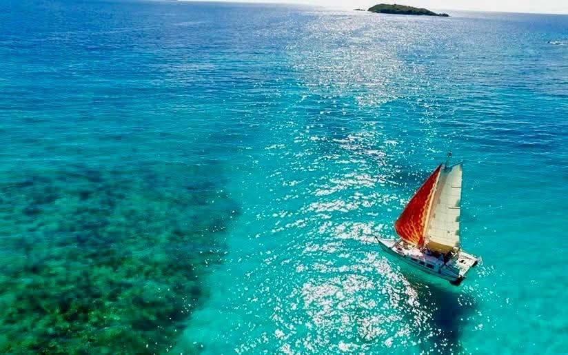 Aerial drone shot of the Mahiya with red sails sailing over a turquoise reef, small VI island in the distance