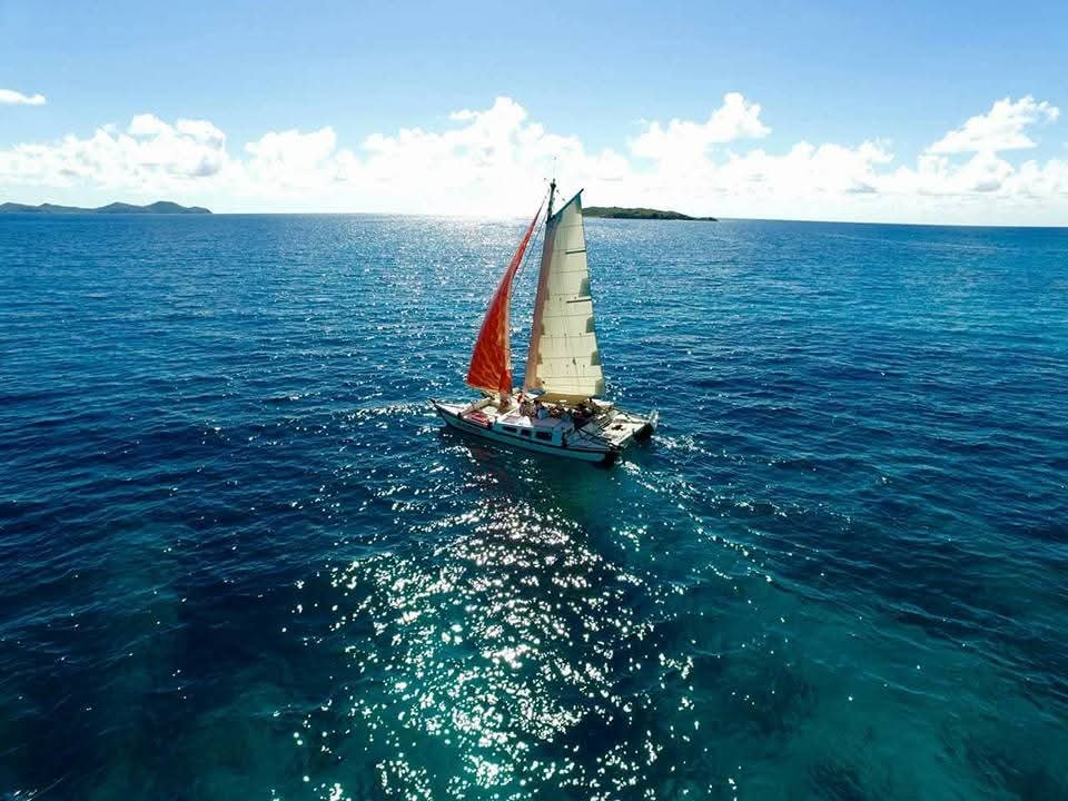 Aerial drone shot of the Mahiya with red sail on sparkling turquoise water, small VI island in the distance