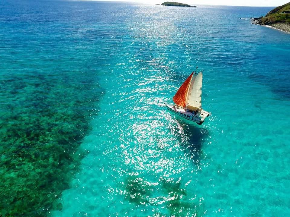 Aerial drone shot of the Mahiya with red sails gliding over brilliant turquoise reef water, rocky VI coastline nearby