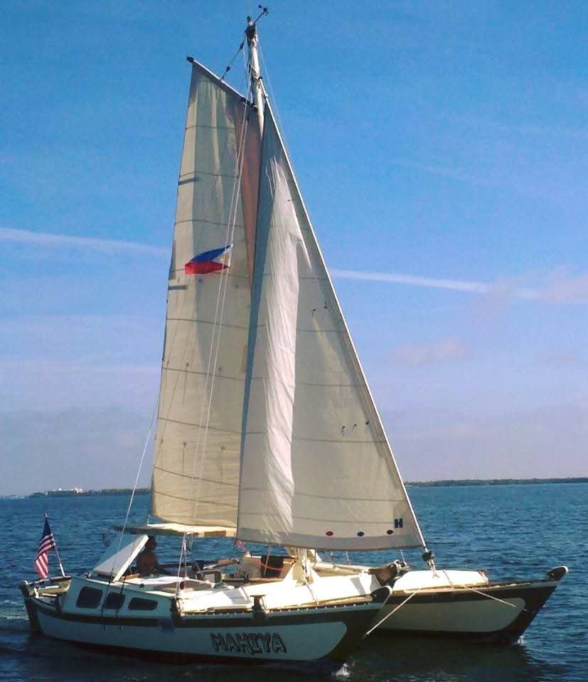 The Mahiya catamaran under full sail on calm water with the American flag flying, boat name on hull