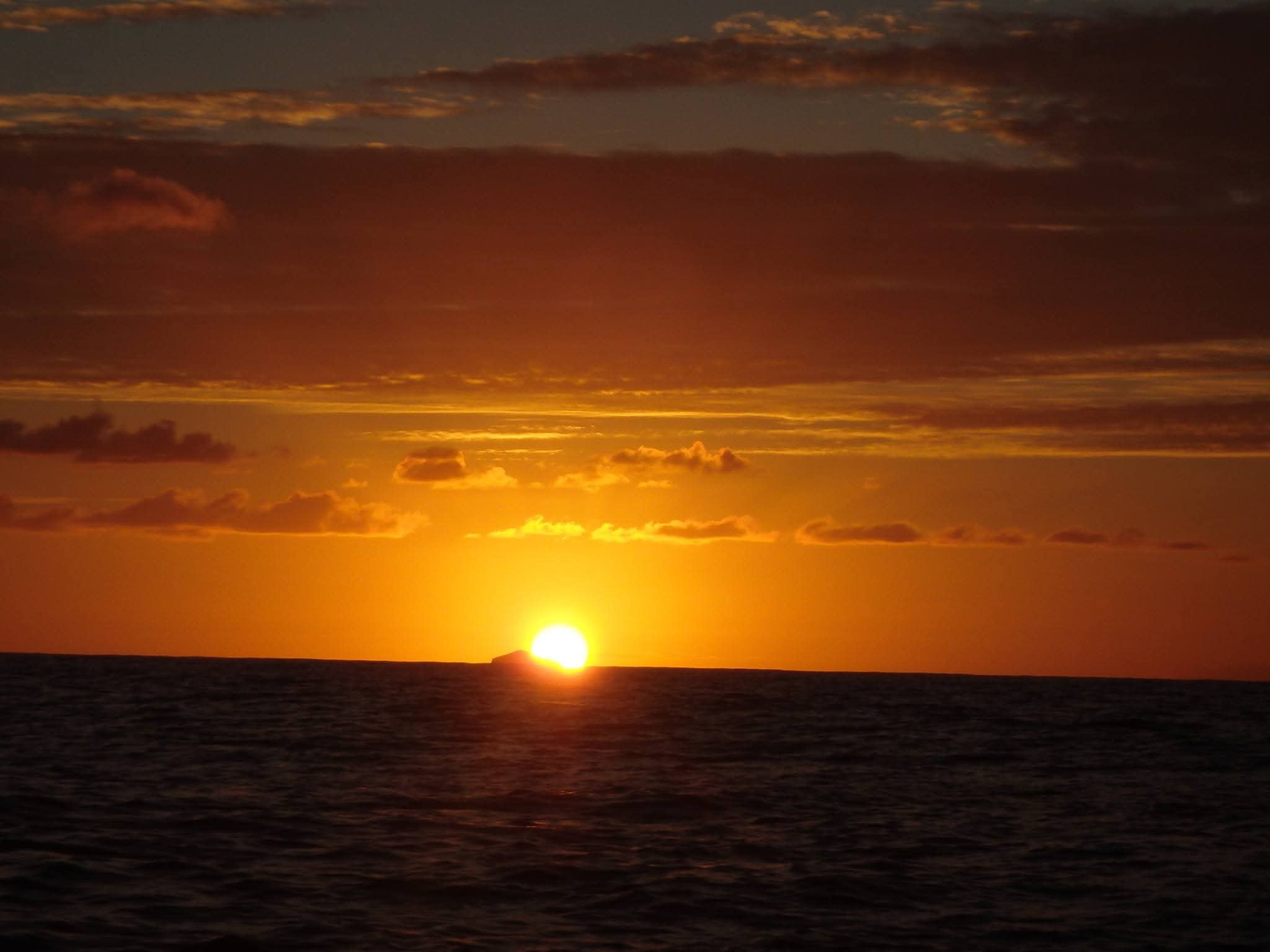Fiery golden sunset over the open ocean with a tiny island silhouetted on the horizon