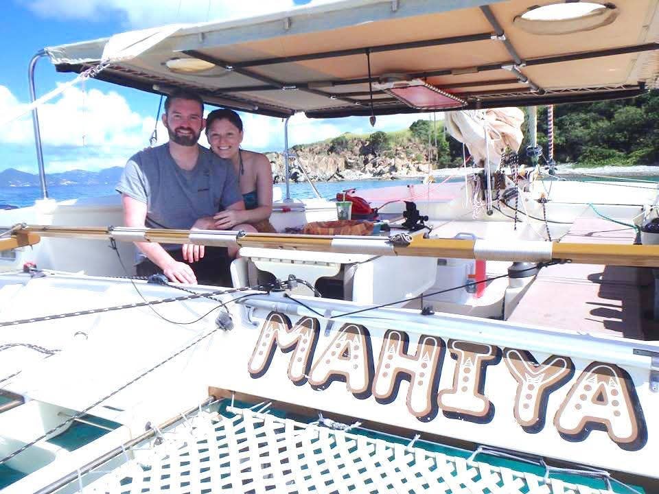 Happy couple smiling aboard the Mahiya catamaran with the boat name visible, rocky VI cove behind