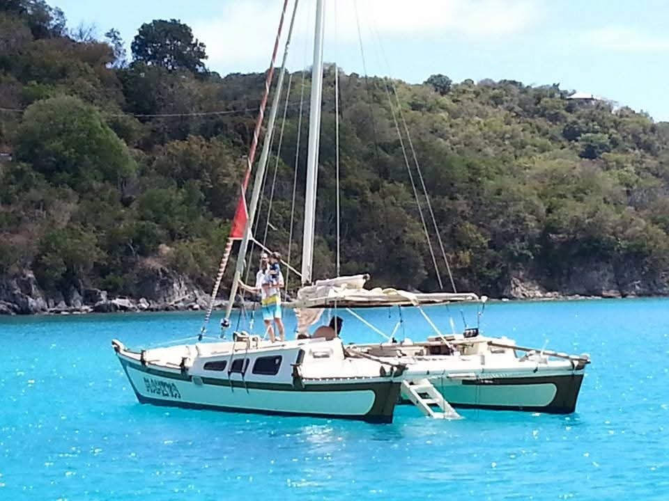 Mahiya catamaran anchored in vivid turquoise water with lush green VI hillside behind, guests on deck