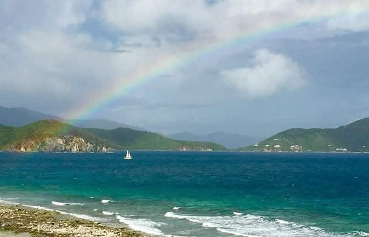 Sailboat on turquoise water beneath a full rainbow arching over green VI mountains and dramatic clouds