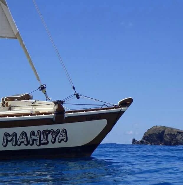 Mahiya bow close-up with MAHIYA name painted on the hull, small rocky VI island in the deep blue background