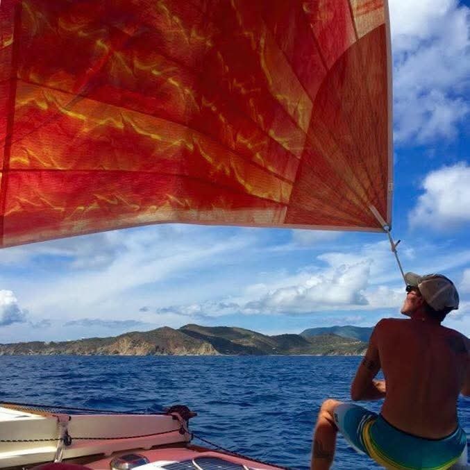 Captain Karl looking up at the vibrant red sail billowing in the wind, lush VI mountains and blue sky behind