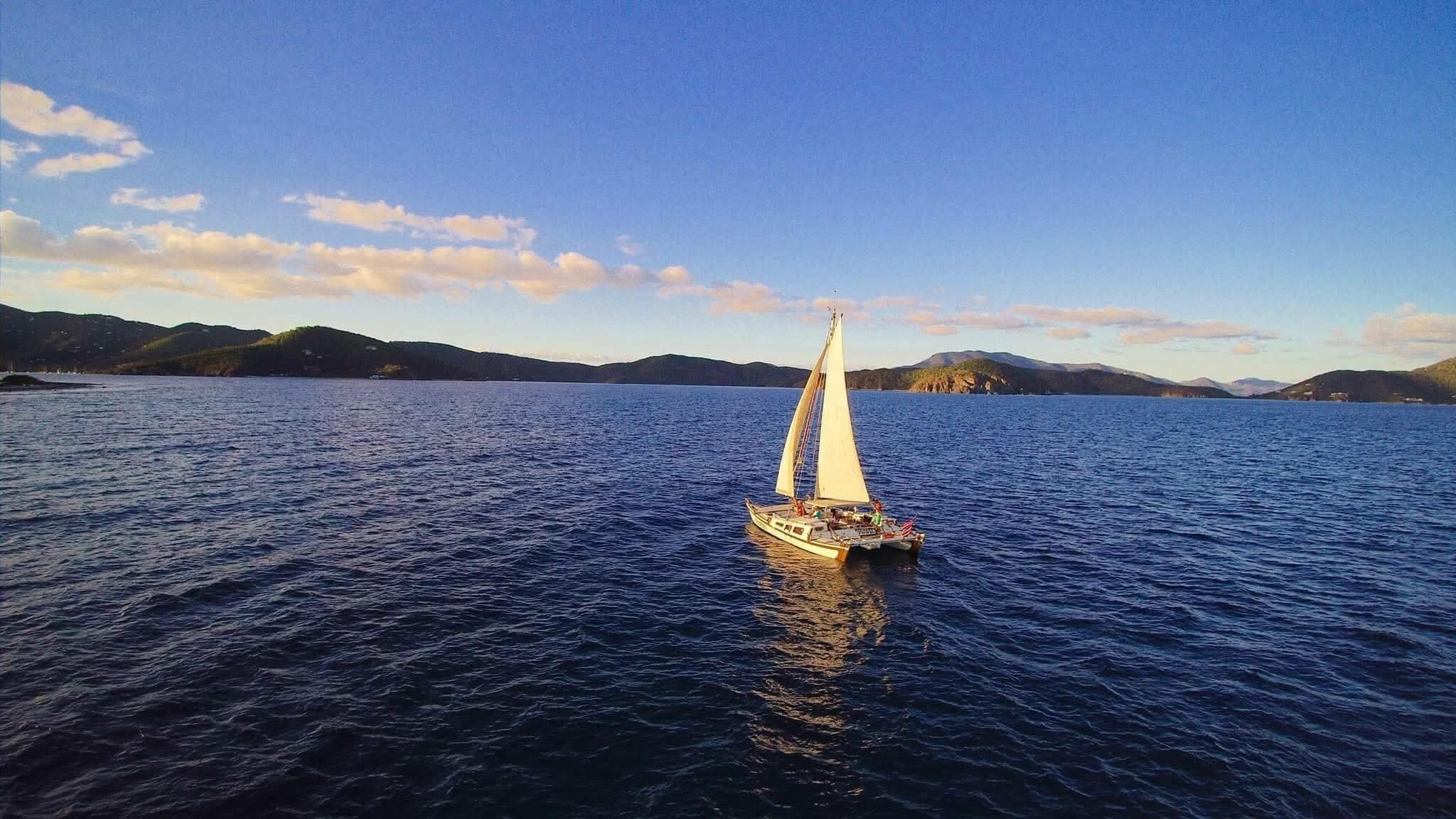 Mahiya catamaran under full white sail on deep blue water with lush VI mountains and golden sky behind