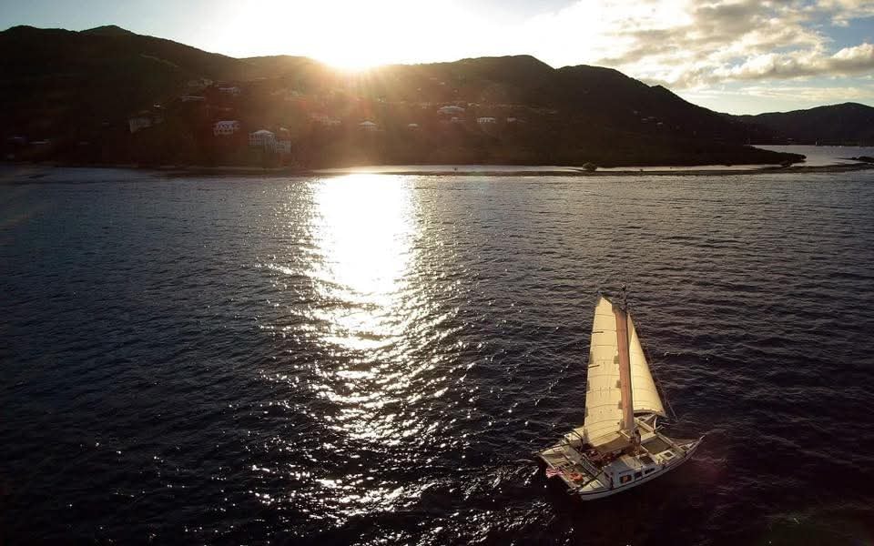 Aerial drone shot of the Mahiya sailing toward a VI town at sunset, golden light reflecting off the water