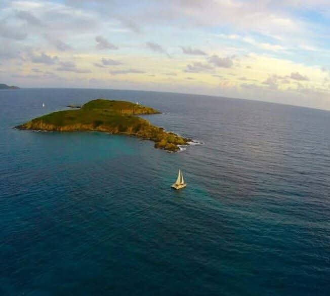 Aerial drone shot of the Mahiya catamaran tiny against a vast teal ocean beside a lush green VI island