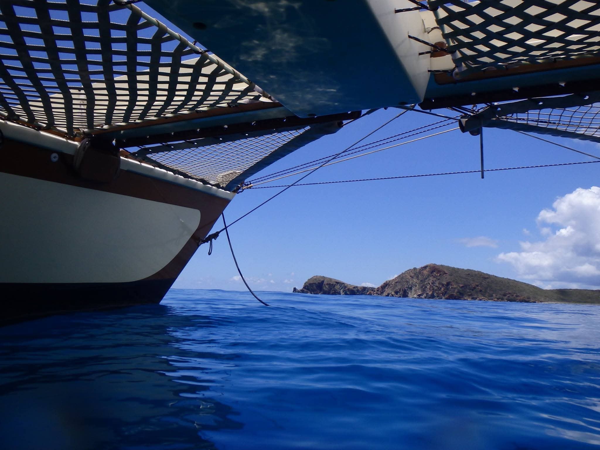 Low water-level view of the Mahiya catamaran hull and bow nets with a rocky VI island ahead under blue sky