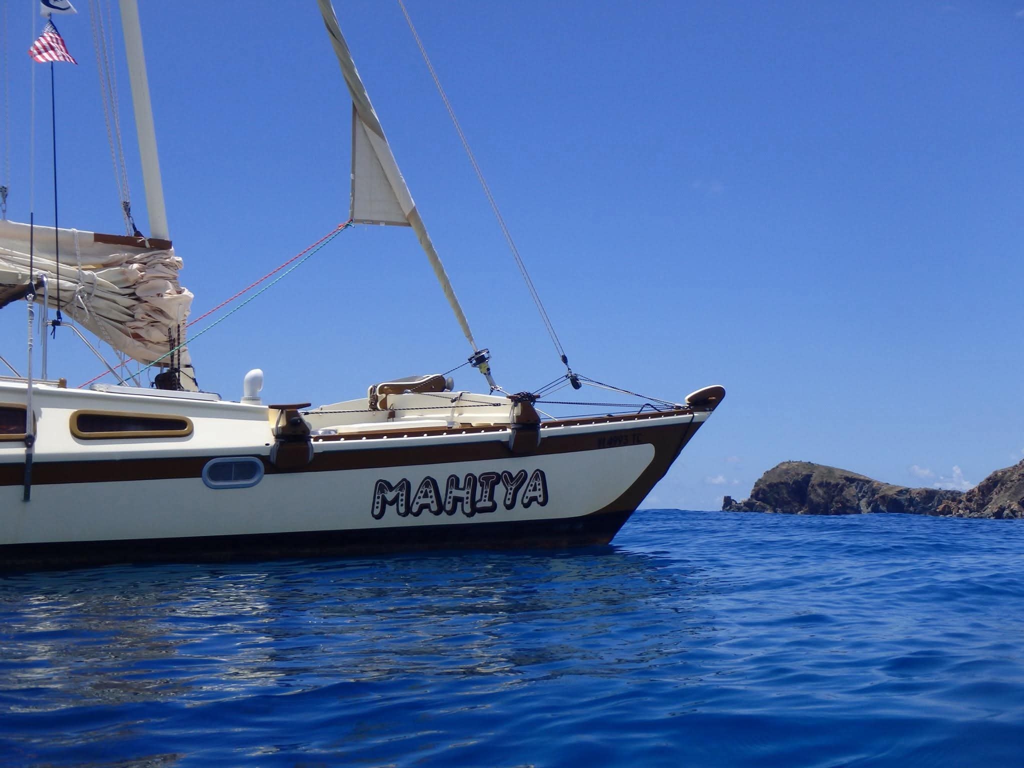 MAHIYA name painted on the hull of the catamaran in decorative lettering, rocky VI island in the background