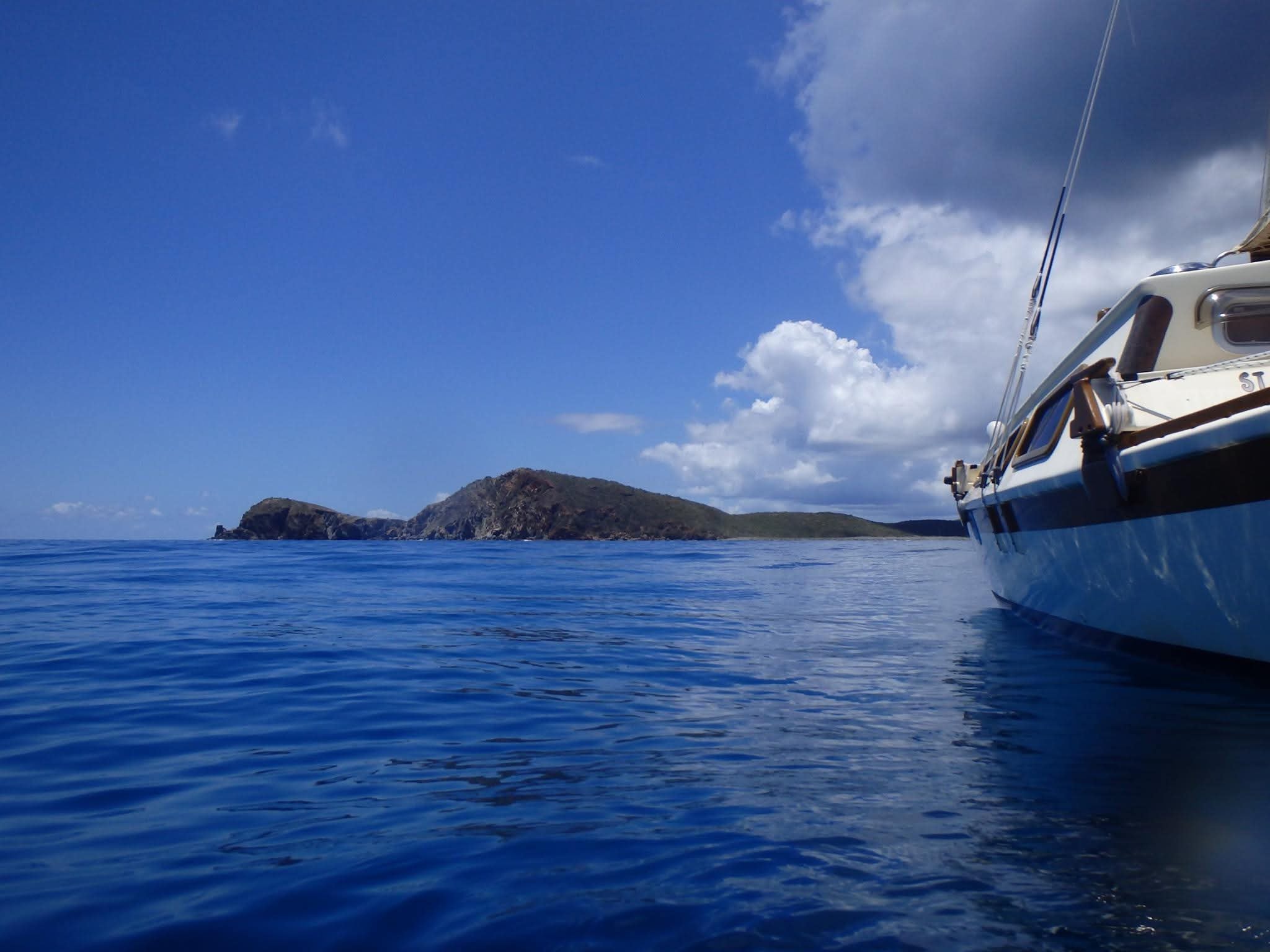 Side view of the Mahiya catamaran hull on calm deep blue water with a dramatic rocky VI island ahead