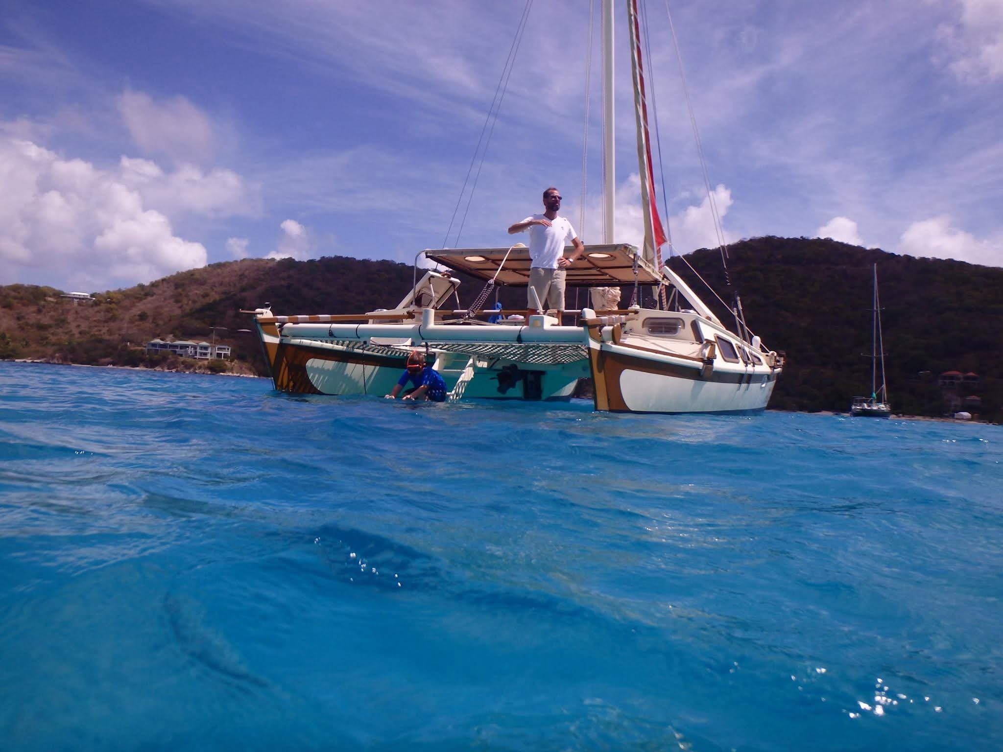 Captain Karl standing on the Mahiya deck waving, guest swimming in brilliant turquoise water below, VI hills behind