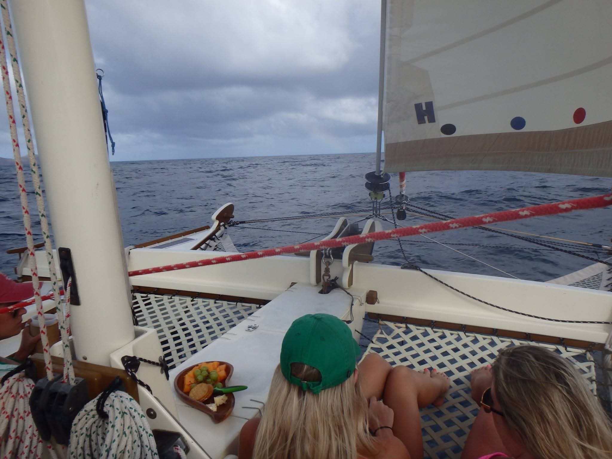 Guests relaxing on the Mahiya catamaran deck with snacks, sails up, open ocean ahead under cloudy skies