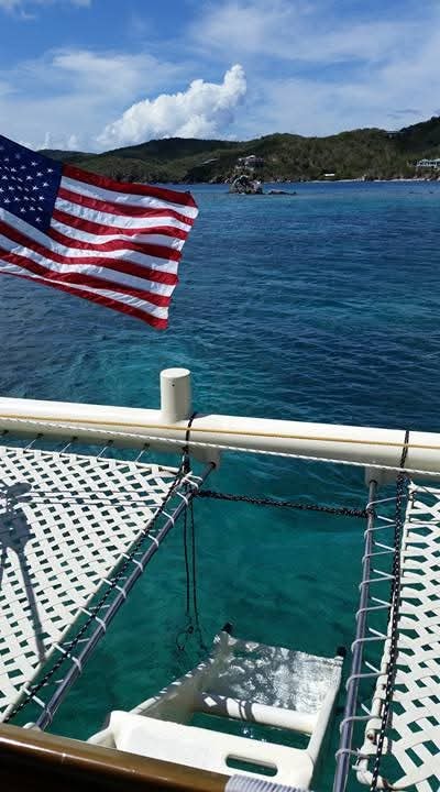 American flag flying from the Mahiya bow net over crystal turquoise water with a rocky VI cove behind