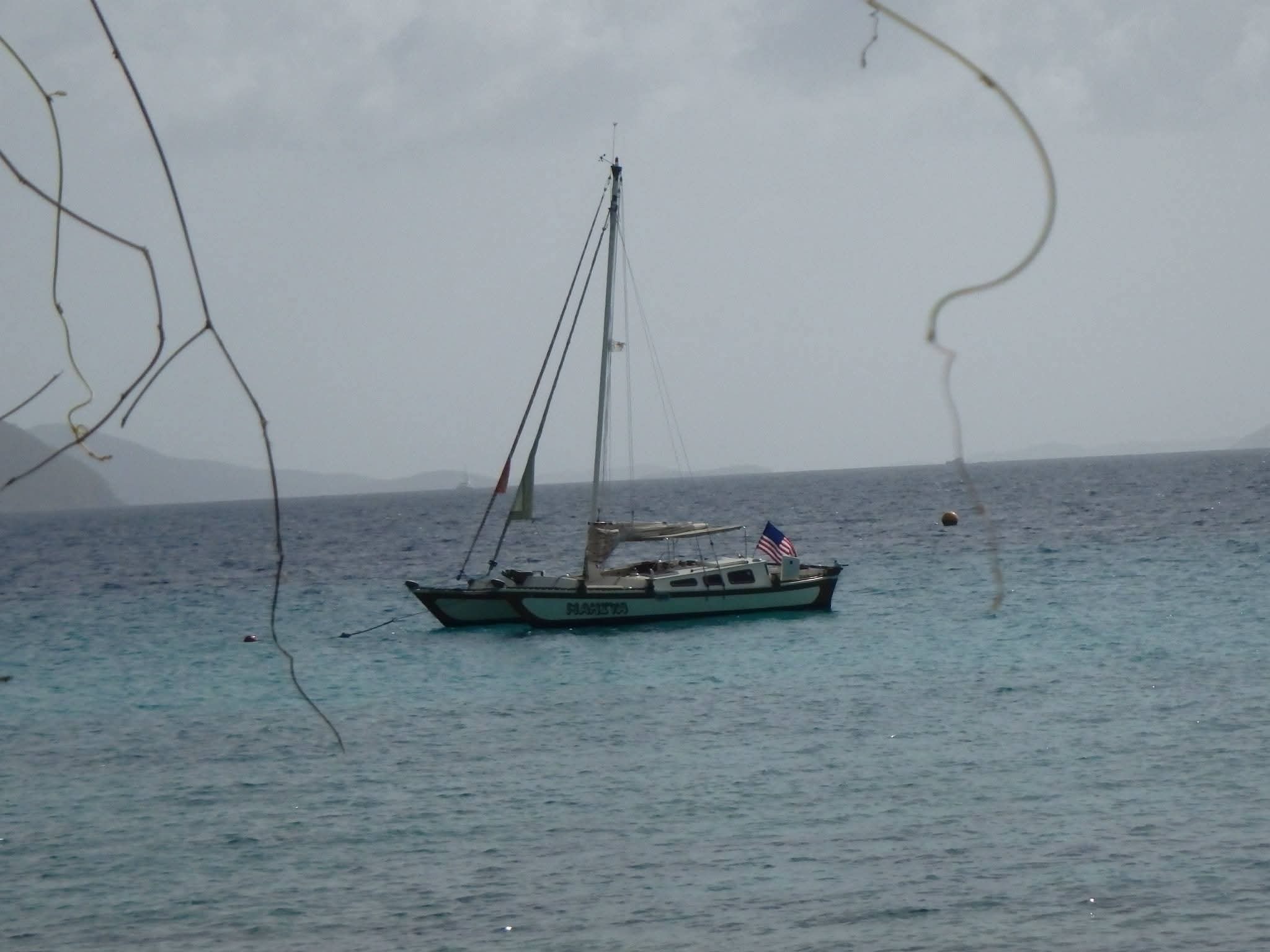 Mahiya catamaran anchored in turquoise water viewed from shore through tropical branches, American flag flying