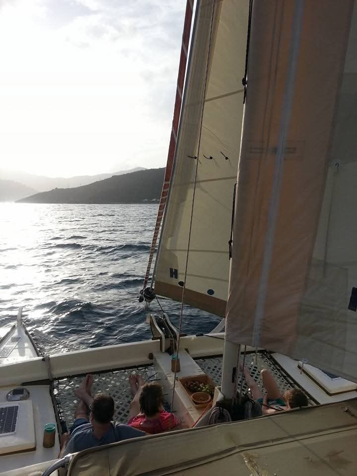 Guests relaxing on the Mahiya deck under full sail, bowl of fruit on deck, VI mountains silhouetted behind